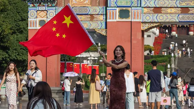 2025年10月7日国庆黄金周假期期间,一位女士在重庆人民大礼堂前挥舞着一面巨大的中国国旗,访客们汇聚于这一地标建筑。广场上遍布爱国装饰和红旗,人们共同庆祝中华人民共和国成立76周年。