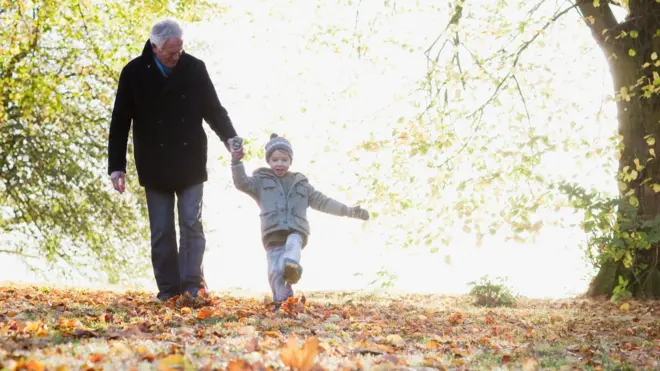 一位年长的绅士与年幼的孙子在秋日落叶中散步,阳光照耀着背景中仍紧贴树枝的绿叶——他们都穿着保暖外套。
