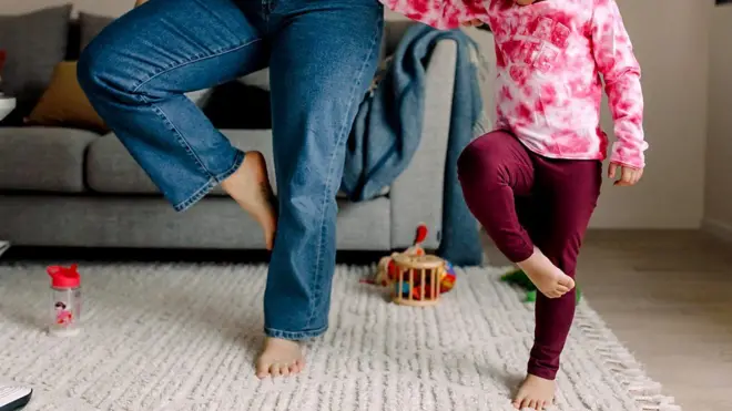 নীল জিন্সের প্যান্ট পরা একজন নারী এবং মেরুন পায়জামা ও সাদা-লাল ছোপ ছোপ টিশার্ট পরা একটি শিশু কার্পেটের ওপর এক পা তুলে দাঁড়িয়ে আছে। তাদের শরীরের ওপরের অংশ ক্যামেরার ফ্রেমের বাইরে। তাদের পেছনে একটি সোফা দেখা যাচ্ছে। কার্পেটে কয়েকটি খেলনা ও একটি ফিডার রাখা আছে