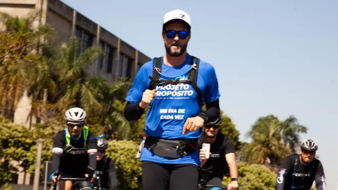 雨果·法里亚斯身穿蓝色跑步上衣,戴着墨镜和帽子跑步。他身后跟着三名骑行者。