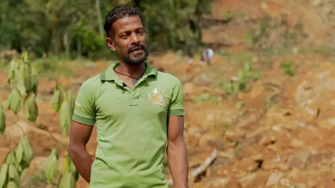 නායයෑමට ලක්වූ ස්ථානයක් පසුබිම්ව සිටින, කොළ පැහැති ටී-ෂර්ට් එකක් ඇඳ සිටින පුද්ගලයෙක්