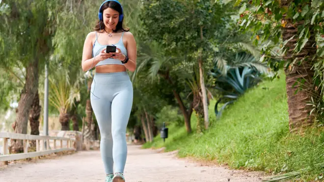 A young woman with dark hair walks along a path while looking at her phone. She wears a blue crop top and leggings, and headphones.