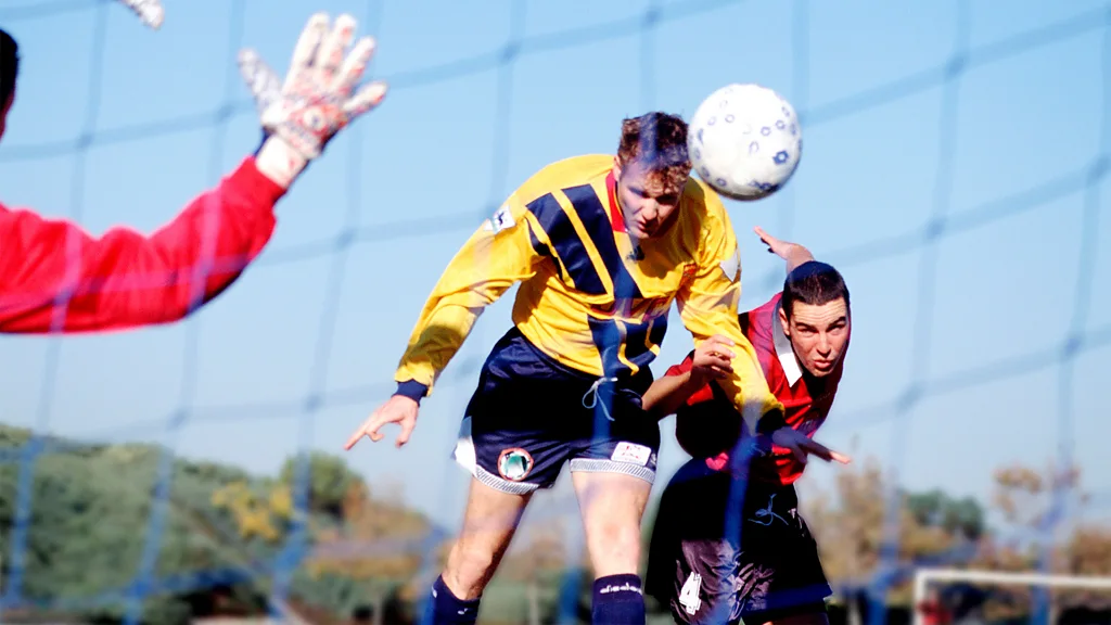 A footballer wearing a yellow jersey with a dark stripe, stretches to head a ball