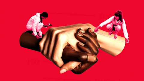 Serenity Strull/ Getty Images Scientists in white protective bodysuits measure and observe two giant hands interlocked in a grip (Credit: Serenity Strull/ Getty Images)