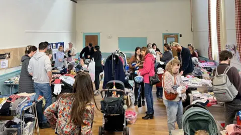 Parents mull around different stalls of clothing inside a village hall.