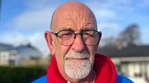 Gordon looking at the camera. He is wearing glasses, a red hoodie and blue coat.