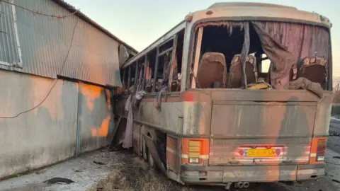 Armed Forces of Ukraine/Telegram The rear end of a burnt and badly damaged bus. It appears to have driven into a wall, with its front end obscured through it. Its windows have been blown out and charred purple curtains hang out of the window frames. Its red and white paint is visible underneath a layer of soot. 