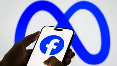 A hand shown holding a smartphone, which is displaying Facebook's blue and white logo on its screen against a larger backdrop of the Meta logo behind it.