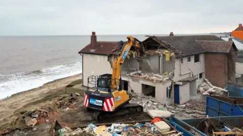 A yellow-framed digger is being used to destroy a house or apartment block. There is debris all around. To the left of the frame is a beach, and in the background is the sea. It is overcast above.