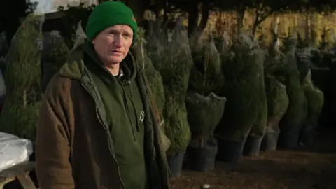 Jonathan Mearns is seen wearing a green beanie and green hoodie stands outdoors beside rows of potted Christmas trees wrapped in netting, with trees and foliage visible in the background.