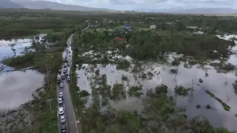 Cars are seen along a road which is surrounded by floodwaters