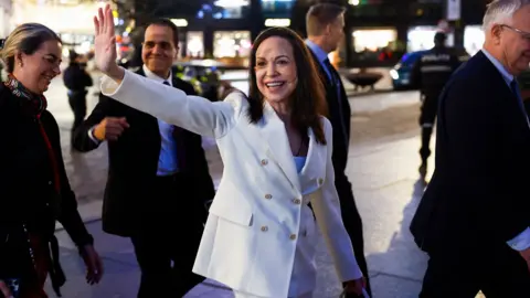 María Corina Machado waving at crowds she is smiling and wearing a white suit