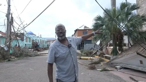 Nick Davis, wearing a short sleeve shirt, points to a destroyed building as a fallen roof is seen in the background