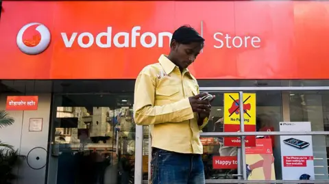 Getty Images A pedestrian wearing a yellow shirt and blue jeans uses his mobile phone outside a Vodafone store in Mumbai
