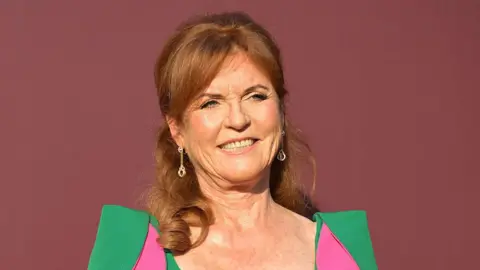 Sarah Ferguson stood in front of a dark red back drop. She is wearing a green dress with pink parts to it. 