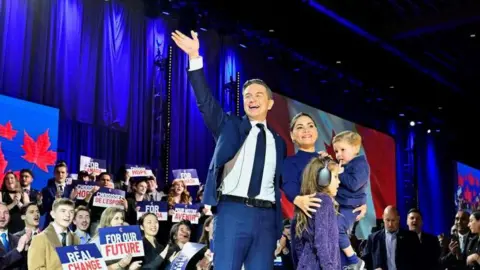 An image of Pierre Poilievre waving to a crowd, next to his wife and two children. He is wearing a a blue suit and tie, and his wife is wearing a dark blue dress and holding their youngest child. Behind him is multiple rows of supporters holding signs that say "REAL CHANGE" and "FOR OUR FUTURE". 