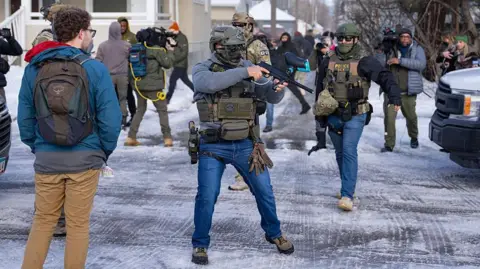 An officer with US Immigration and Customs Enforcement or ICE (right) and another federal officer holding a crowd control device (centre) stand at a Minneapolis intersection where protesters had gathered after the death of Renee Nicole Good. Bystanders and journalists also stand in the intersection.