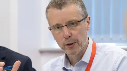 Peak Cluster Chief Executive John Egan, who has short grey hair, rimless glasses and who is wearing a light blue shirt with an orange lanyard, pictured mid sentence sat at a table next to a man in a blue suit jacket. 