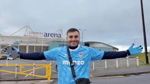 man with brown hair stood outside the Coventry Building Society Arena near a yellow car park barrier with his arms outstretched. The man is wearing a blue replica Coventry City football shirt with a long-sleeved navy t-shirt underneath with blue gloves with navy fingertips.