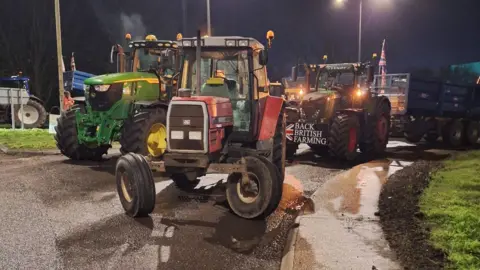 Multiple tractors are parked on a road, completely blocking access. One has a sign that says "Back British farming" on the front.