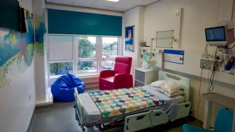 A hospital room with a bright coloured mural of a beach/palm tree scene, a blue bean bag, a pink chair and a patterned, multicoloured blanket over the hospital bed.