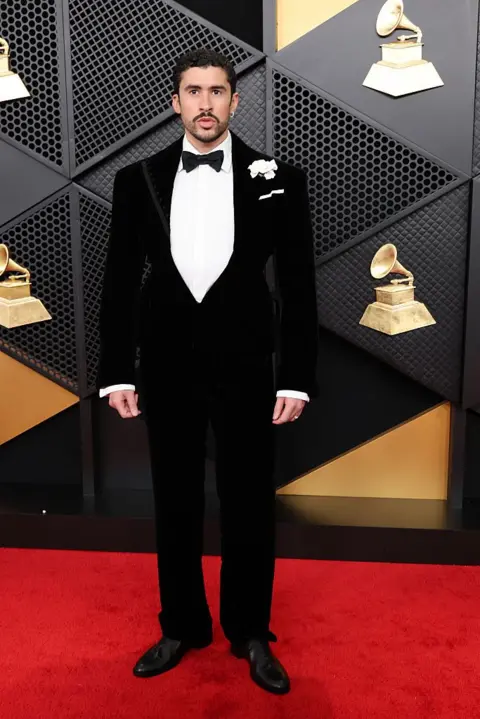 Getty Images Bad Bunny wears a tuxedo with a white flower pinned near his left shoulder as he posed for a photo on the Grammy Award red carpet.