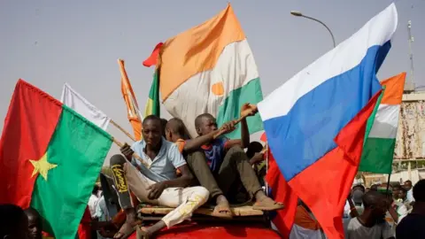 Protesters display Russia's and Niger's flags as they sit on top of a vehicle amid a demonstration.
