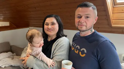 Holly and Dean stood next to each other, Holly is smiling and holding their baby. Holly has short black hair and Dean has shorter dark hair. He's holding a mug. 