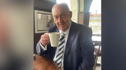 A man with grey hair wearing a blazer, tie and white shirt sitting at a table  while holding up a coffee mug.