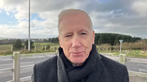 Campbell is looking right at the camera. He has short white hair and is wearing a dark grey coat and has a black scarf tucked in. 