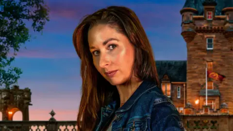A close-up image of Rachel Duffy from County Down. She is looking at the camera with a neutral expression. She has long, dark hair and is wearing a blue denim jacket. Behind her is the traitors Castle, with the sky getting darker as the sun sets.