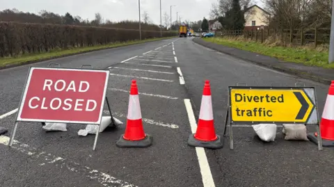 Signs and traffic cones placed on a road. One sign reads 'Road Closed' and the other is an arrow pointing right for 'Diverted traffic. In the distance there is a lorry and flashing flue lights.