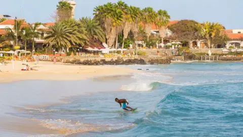 A man surfing near the coastline of  Santa Maria, Sal Island, Cape Verde. The water is a pale blue and he is riding a small wave. There are palm trees and  white buildings with terracotta-coloured roofs in the background.