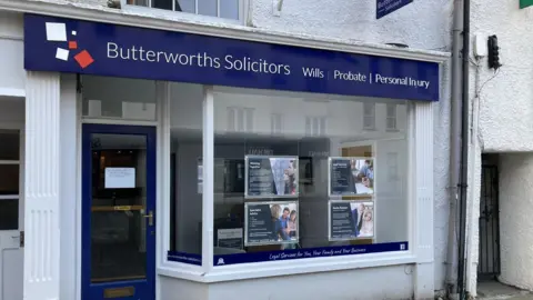 The front of a solicitors office. There is a big glass window with some advertising panels hung in it. There is a blue sign above the whole shop frontage which says 'Butterworths Solicitors.' The door is glass with a blue trim. In the centre of the door is a white piece of paper stuck to it with writing on it.