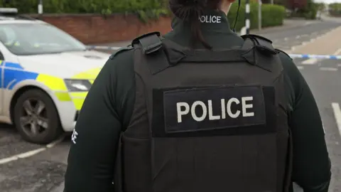 A police officer in a bottle green uniform with a black vest on with 'police' written across it in white writing stands in the middle of the road. A police car sits parked to their left. 