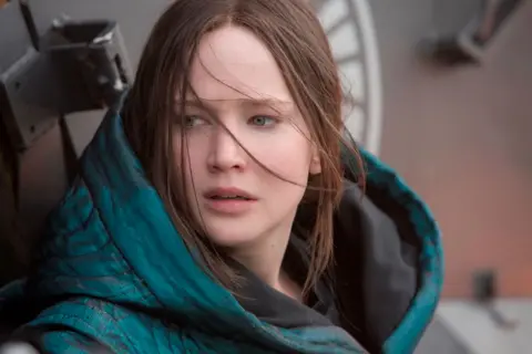 A still from the Hunger Games shows Jennifer Lawrence's character wearing a green, hooded coat. She's looking off to the side, the wind sweeping fronds of hair across her face, at an unseen threat.