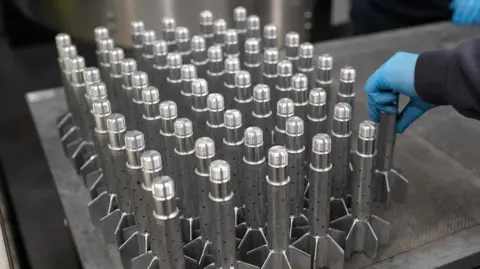 The tail sections of around 60 mortar munitions are lined up on a table. They look like small rockets. 