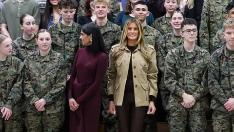 Usha Vance wears a burgundy dress and stands next to Melania Trump in a khaki jacket as they pose for a photo surrounded by high school students dressed in military uniforms.
