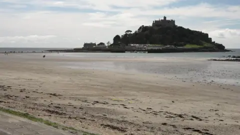 A view of St Michael's Mount from Marazion. The beach is sandy and the tide is out. The island has a castle situated at the top.