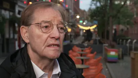 Pat Karney stands in front of a bar on Manchester's Canal Street. He has short hair and wears round glasses. He wears a black coat over a white shirt