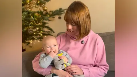 Jessica sits on a sofa, holding her baby son in her arms. She is wearing a pastel pink hoodie, while the baby is dressed in a light grey outfit and gripping a small yellow toy. In the background, a decorated Christmas tree with lights and ornaments is visible.
