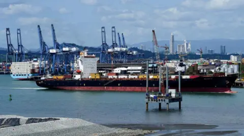A large black and red container ship sails near the Balboa Port, as other container ships and cranes are seen in the background