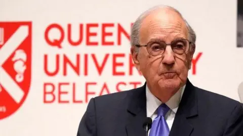 George Mitchell, with short grey hair, wearing glasses, a navy suit, white shirt and purple tie. He is standing behind a white podium. There is a white wall behind him with Queen's University Belfast and the university logo in red. 