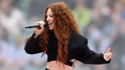 Jess Glynne is singing into a mic, she is wearing a cropped black suit jacket. SHe has long curly ginger hair. 