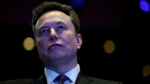 A close-up shot of Elon Musk in a dark room. He stands with a neutral expression against a black background. He has short dark brown hair, and wears a black jacket, white shir,t and dark tie.