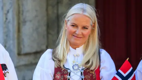 Per Ole Hagen/Getty Images Crown Princess Mette-Marit, in traditional attire, smiling and holding a small Norwegian flag