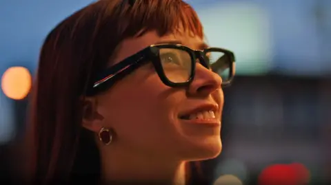 A close-up of a woman wearing the glasses. She is smiling and has long red hair. The glasses are black with thick rims, and the lenses seem slightly translucent.