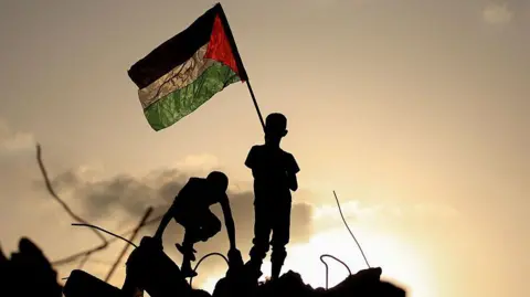 Palestinian children play as one waves a Palestinian national flag as he stands on the rubble of a destroyed building at the Bureij camp in Gaza. They are silhouetted against a setting sun illuminates the background of the image. 