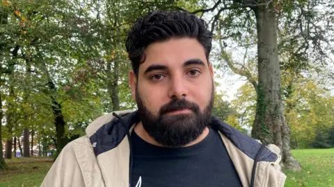 A man with dark hair and a beard is pictured in a park area. He is wearing a dark-coloured t-shirt and a cream coat. Behind him is a grassy area with trees. 