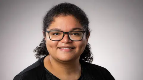 A professional headshot of Dr Nina Higson-Sweeney wearing a black t shirt and black rectanglar framed glasses 